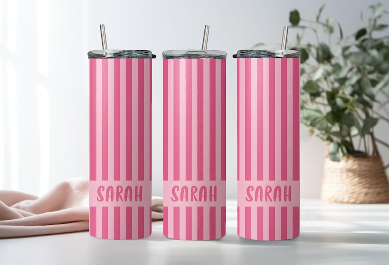 May include: Three tall, cylindrical stainless steel tumblers featuring a vertical pink and light pink striped pattern. Each tumbler has a clear lid with a metal straw and a solid pink horizontal band near the base with the name Sarah in pink.