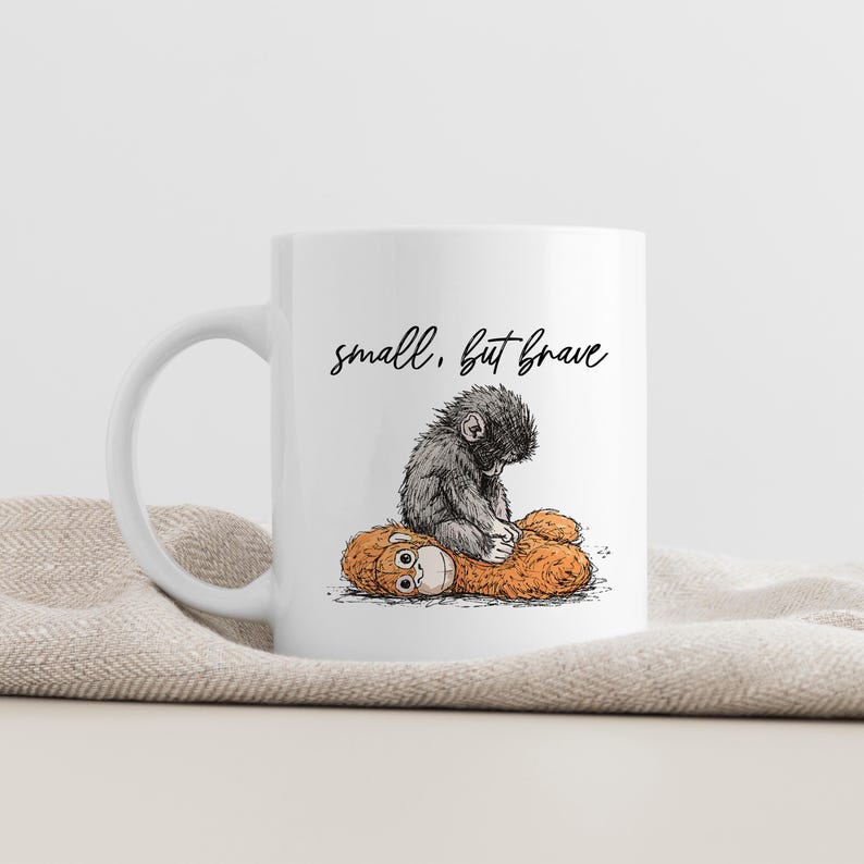 May include: White ceramic mug featuring a whimsical illustration of a small, gray monkey sitting atop a larger, orange plush toy. The mug has the text "small, but brave" in a handwritten font.