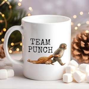 May include: White ceramic mug with the text "TEAM PUNCH" and a cartoon illustration of two monkeys. One monkey is pulling the other. Marshmallows and pinecones are in the background.