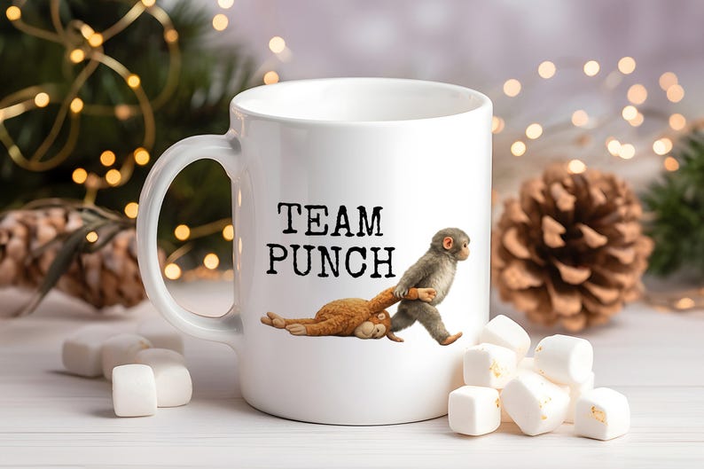May include: White ceramic mug with the text "TEAM PUNCH" and a cartoon illustration of two monkeys. One monkey is pulling the other. Marshmallows and pinecones are in the background.