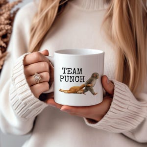May include: White ceramic mug with the text "TEAM PUNCH" and a cartoon illustration of two monkeys. One monkey is pulling the other. The mug is held by a person wearing a white sweater.