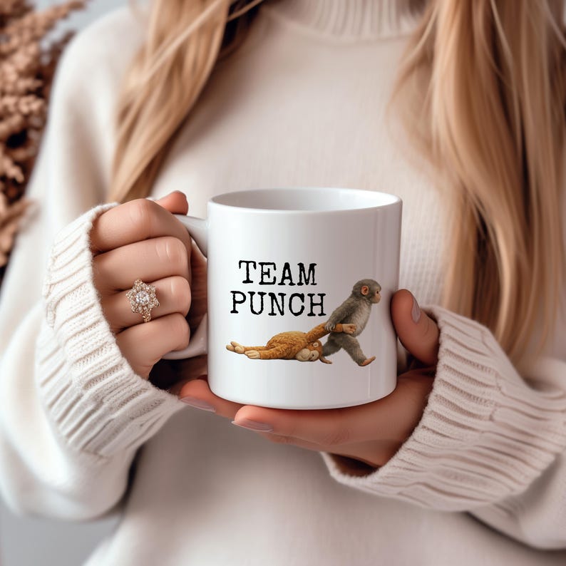 May include: White ceramic mug with the text "TEAM PUNCH" and a cartoon illustration of two monkeys. One monkey is pulling the other. The mug is held by a person wearing a white sweater.