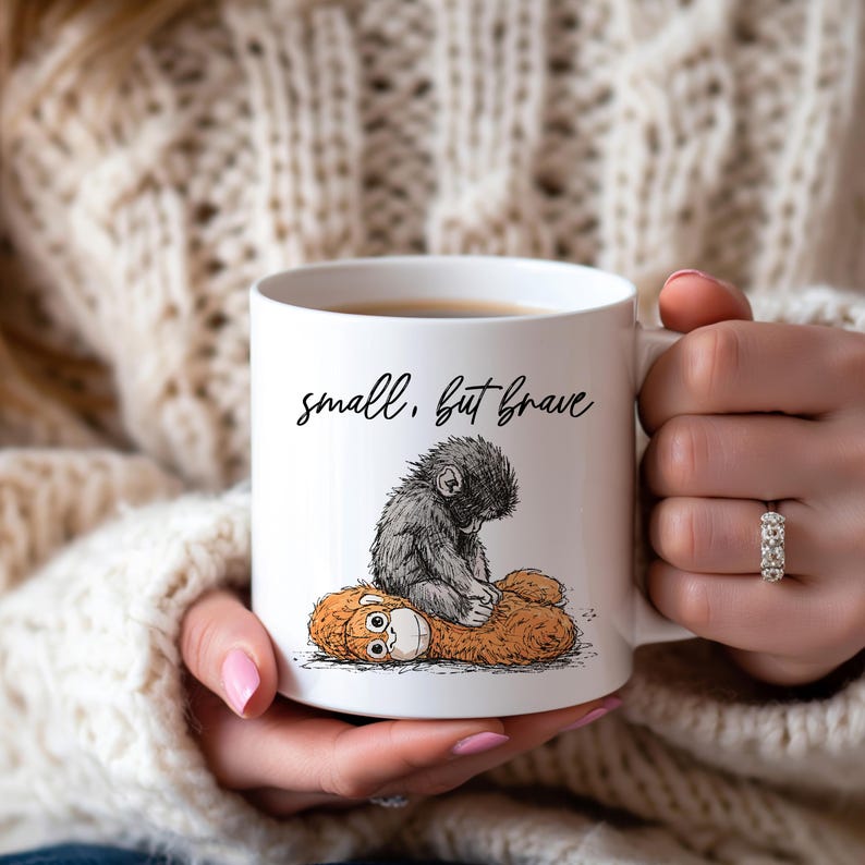 May include: White ceramic mug with the text "small, but brave" in black script. The mug features a black and gray illustration of a small monkey sitting on a brown stuffed animal.