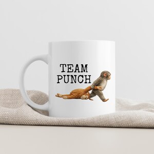 May include: White ceramic mug with the text "TEAM PUNCH" in black. The mug features a graphic of two monkeys, one pulling the other. The background is a neutral color.