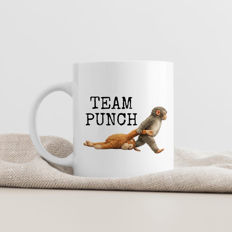 May include: White ceramic mug with the text "TEAM PUNCH" in black. The mug features a graphic of two monkeys, one pulling the other. The background is a neutral color.
