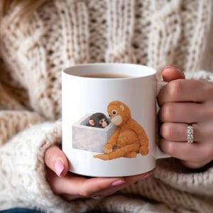 May include: White ceramic mug featuring an illustration of two cartoon orangutans. One orangutan is sitting, and the other is peeking out of a gray box. The mug is filled with a dark liquid.