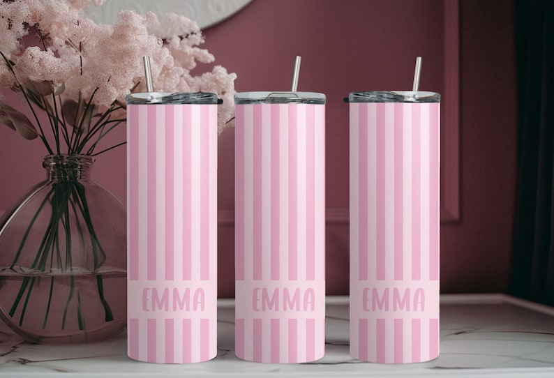 May include: Three cylindrical stainless steel tumblers with lids and straws, featuring a vertical light pink and white striped pattern. Each tumbler has a solid pink horizontal band near the base with the name EMMA printed in a light pink font.