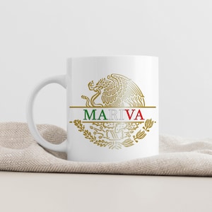 May include: White ceramic mug featuring a gold emblem with an eagle and the text "MARIVA" in green and red. The mug has a curved handle and is set against a neutral background.
