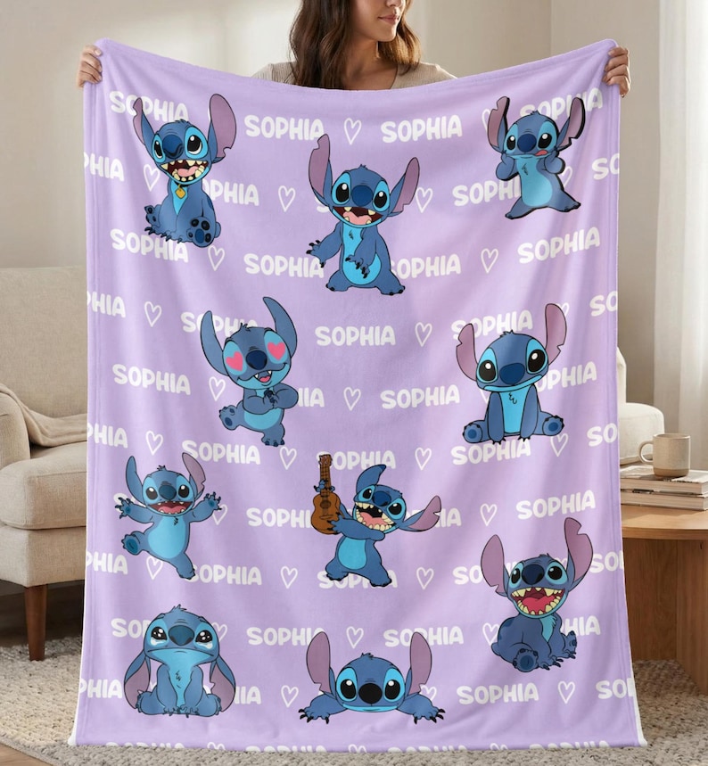 May include: A lavender blanket featuring multiple illustrations of a blue cartoon character. The word "SOPHIA" and heart symbols are printed across the blanket. The blanket is held up by a person.