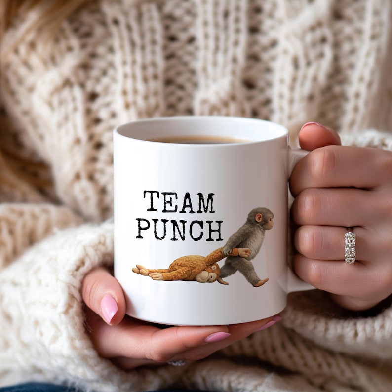 May include: White ceramic mug with the text "TEAM PUNCH" in black. The mug features a cartoon illustration of a monkey pulling another monkey by a stick. The mug is filled with a dark liquid.
