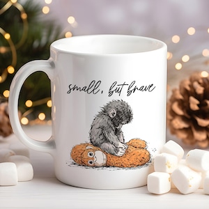 May include: White ceramic mug with a whimsical illustration of a small, gray creature sitting on a larger, orange creature. The text "small, but brave" is written above the illustration. Marshmallows and pine cones are in the background.
