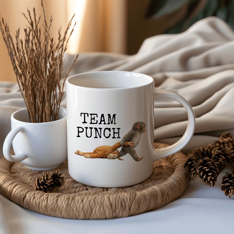 May include: White ceramic mug with the text "TEAM PUNCH" and an illustration of a monkey pulling a cat. A smaller white mug and decorative items are in the background. The mug is on a woven tray.