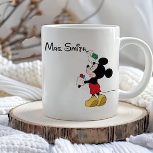 May include: White ceramic mug featuring a cartoon Mickey Mouse character holding markers and a green bottle, with the text "Mrs. Smith" in black script. The mug is on a wooden coaster.