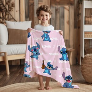 May include: A light pink blanket featuring multiple blue cartoon character illustrations. The blanket has the name "SOPHIA" and heart shapes printed across it. The blanket is held up by a child.