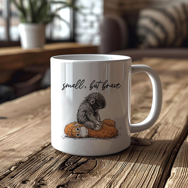 May include: White ceramic mug featuring a whimsical illustration of a small, gray monkey sitting on a plush toy. The mug has the text "small, but brave" in a handwritten font. The mug is on a wooden surface.