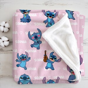 May include: A pink blanket featuring cartoon character Stitch in various poses, with the name "SOPHIA" and heart graphics. The blanket has a white, plush underside, and is folded. The blanket is made of a soft material.