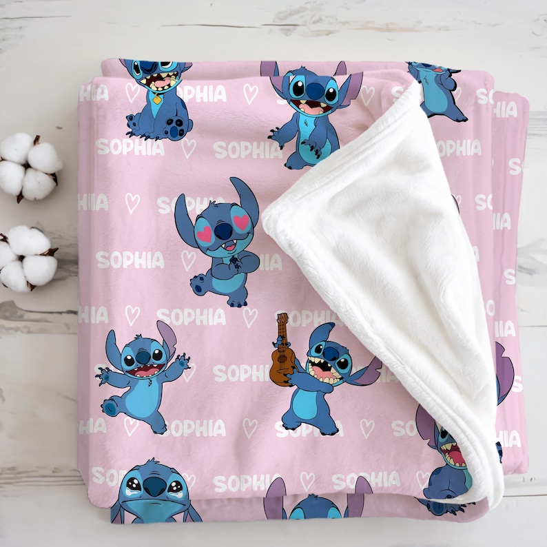 May include: A pink blanket featuring cartoon character Stitch in various poses, with the name "SOPHIA" and heart graphics. The blanket has a white, plush underside, and is folded. The blanket is made of a soft material.