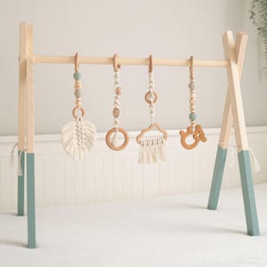 Può includere: Un palazzetto per giochi per bambini in legno con cinque giocattoli appesi. I giocattoli includono un anello di legno, un giocattolo a forma di nuvola con nappe, un giocattolo a forma di foglia con nappe, un orso di legno e un giocattolo con perline di legno.