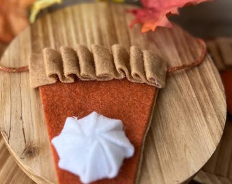 May include: A felt pumpkin pie slice with a dollop of white whipped cream, part of a fall-themed banner. The pie is orange with a brown crust. Decorative fall leaves in shades of yellow, orange, and red surround the pie.