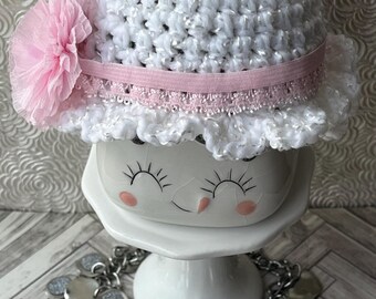 May include: A white crocheted baby hat with a pink flower and a pink elastic band. The hat has a ruffled brim and is displayed on a white ceramic stand. A silver necklace with round, glittery charms is in the foreground.