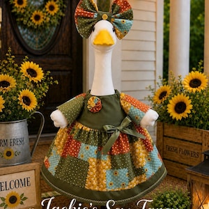 May include: A white goose figurine wearing a patchwork dress with olive green, orange, and light blue floral patterns, a matching oversized bow on its head, and a small fabric flower accent on the bodice, featuring a bright yellow beak.