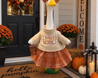 May include: A white goose figurine wearing a tan turtleneck sweater with the text feelin' pumpkin spicy, an orange plaid skirt, and a matching orange plaid bow on its head. The goose holds a small brown cup labeled pumpkin spice.