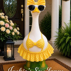 May include: A white goose statue wearing a yellow and white polka dot bikini top, a matching ruffled skirt, a large bow on its head, and white-framed sunglasses. The base is green, and the text Jackie's Sew Fancy is written in white script.