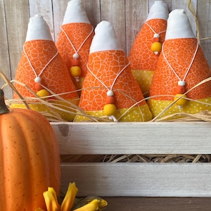 May include: A wooden crate filled with decorative candy corn plushies. Each plushie has an orange patterned body, a white top, and a yellow base. They are adorned with small beaded necklaces. A pumpkin and a sunflower are in the foreground.