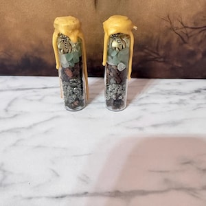 May include: Two clear glass vials sealed with yellow wax dripping down the sides. Inside each vial are layers of small green stones, dark brown pieces, and metallic pyrite chunks. A small bronze-colored charm shaped like a money bag is attached.