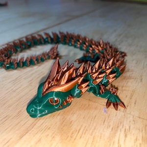 Puede incluir: Una figura de dragón impresa en 3D de color verde y cobre con púas a lo largo de la espalda y la cola. El dragón tiene una cabeza detallada con ojos grandes y una cresta espinosa.