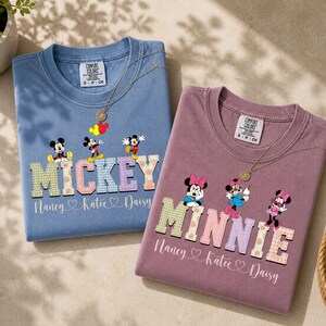 Könnte beinhalten: Zwei T-Shirts, eines in Blau und eines in Mauve, jeweils mit einer goldenen Halskette. Das blaue Shirt zeigt Micky Maus und das Wort "MICKEY" in einer karierten Schriftart. Das mauvefarbene Shirt zeigt Minnie Maus und das Wort "MINNIE".