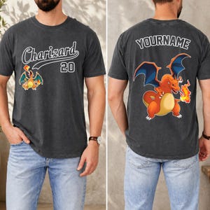 May include: Dark grey t-shirt with a cartoon Charizard graphic and the text "Charizard 20" on the front. The back of the shirt features a larger Charizard design and the text "YOURNAME". The shirt is worn by a person.