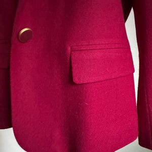 Pu&ograve; includere: Primo piano di un blazer magenta con un bottone dorato e una tasca. Il blazer &egrave; realizzato in un tessuto morbido e strutturato. Il design del blazer &egrave; classico e su misura.