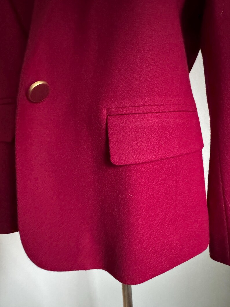 Pu&ograve; includere: Primo piano di un blazer magenta con un bottone dorato e una tasca. Il blazer &egrave; realizzato in un tessuto morbido e strutturato. Il design del blazer &egrave; classico e su misura.