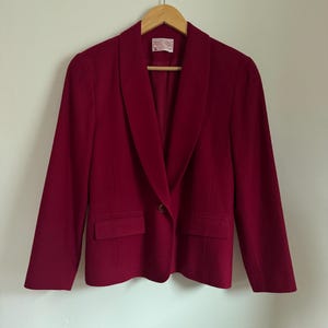 Pu&ograve; includere: Blazer bordeaux con collo a scialle, due tasche frontali e chiusura con un solo bottone. La giacca &egrave; appesa a una gruccia di legno su uno sfondo bianco. Il tessuto del blazer &egrave; di colore uniforme.