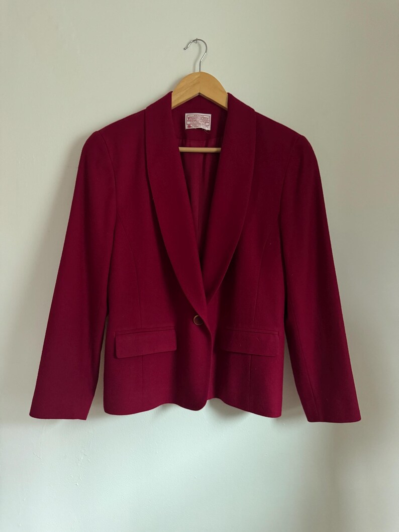 Pu&ograve; includere: Blazer bordeaux con collo a scialle, due tasche frontali e chiusura con un solo bottone. La giacca &egrave; appesa a una gruccia di legno su uno sfondo bianco. Il tessuto del blazer &egrave; di colore uniforme.