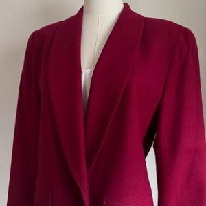 Pu&ograve; includere: Un blazer rosso intenso con un ampio colletto a revers e due tasche frontali. La giacca a maniche lunghe &egrave; esposta su un manichino. Il blazer &egrave; realizzato in un tessuto morbido e strutturato.