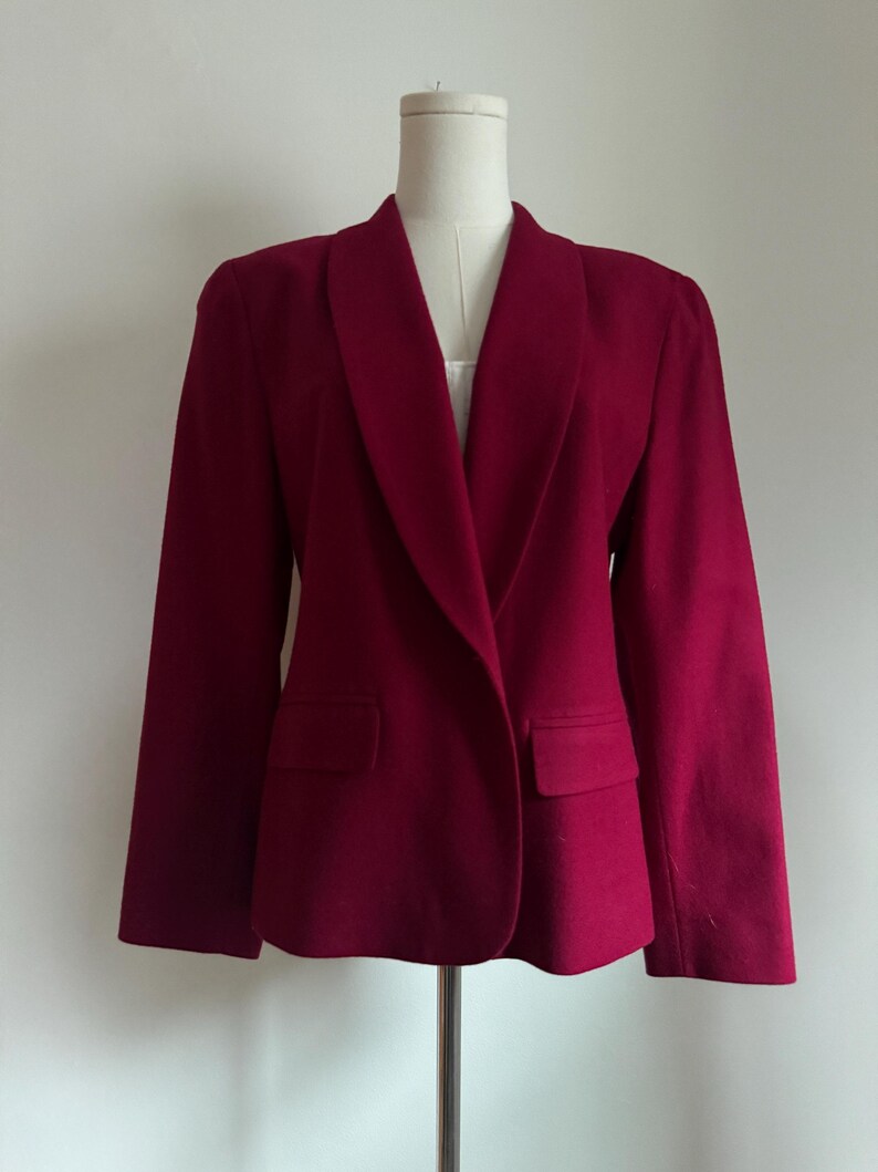 Pu&ograve; includere: Un blazer bordeaux con collo a scialle e due tasche anteriori. La giacca &egrave; esposta su un manichino su uno sfondo bianco. Il colore del blazer &egrave; una tonalit&agrave; ricca e satura e il tessuto sembra essere lana morbida o simile.