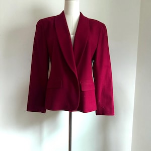 May include: A burgundy blazer with a shawl collar and two front pockets. The jacket is displayed on a mannequin. The blazer has long sleeves and a tailored fit. The fabric appears to be a medium-weight material.