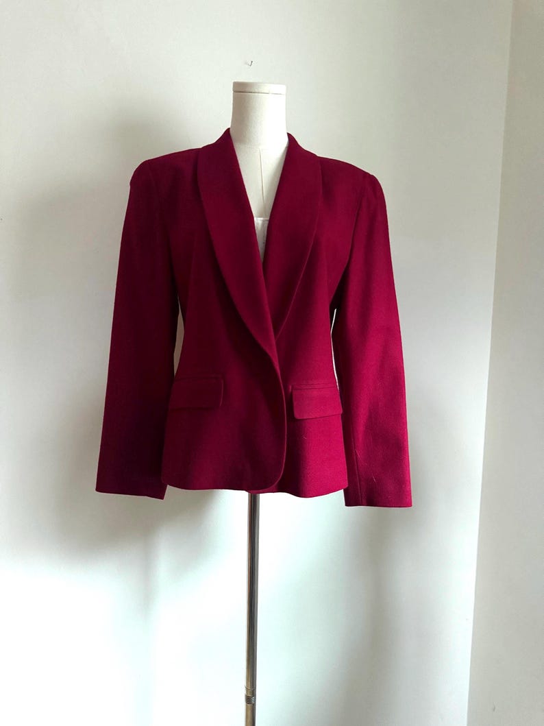 Pu&ograve; includere: Un blazer bordeaux con collo a scialle e due tasche anteriori. La giacca &egrave; esposta su un manichino. Il blazer ha maniche lunghe e una vestibilit&agrave; su misura. Il tessuto sembra essere un materiale di peso medio.