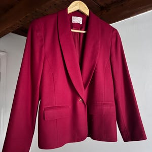 Pu&ograve; includere: Un blazer bordeaux con collo a scialle, due tasche frontali e una chiusura con un unico bottone dorato. Il blazer &egrave; appeso a una gruccia di legno. Il capo sembra essere un misto lana.
