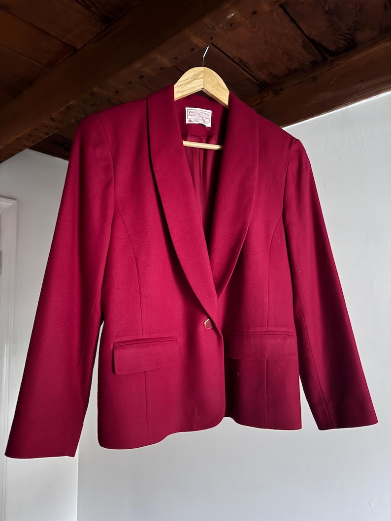 Pu&ograve; includere: Un blazer bordeaux con collo a scialle, due tasche frontali e una chiusura con un unico bottone dorato. Il blazer &egrave; appeso a una gruccia di legno. Il capo sembra essere un misto lana.