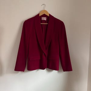 Pu&ograve; includere: Un blazer bordeaux con chiusura a un bottone e due tasche frontali. La giacca &egrave; appesa a una gruccia di legno contro un muro bianco. Il blazer ha un classico design a revers.