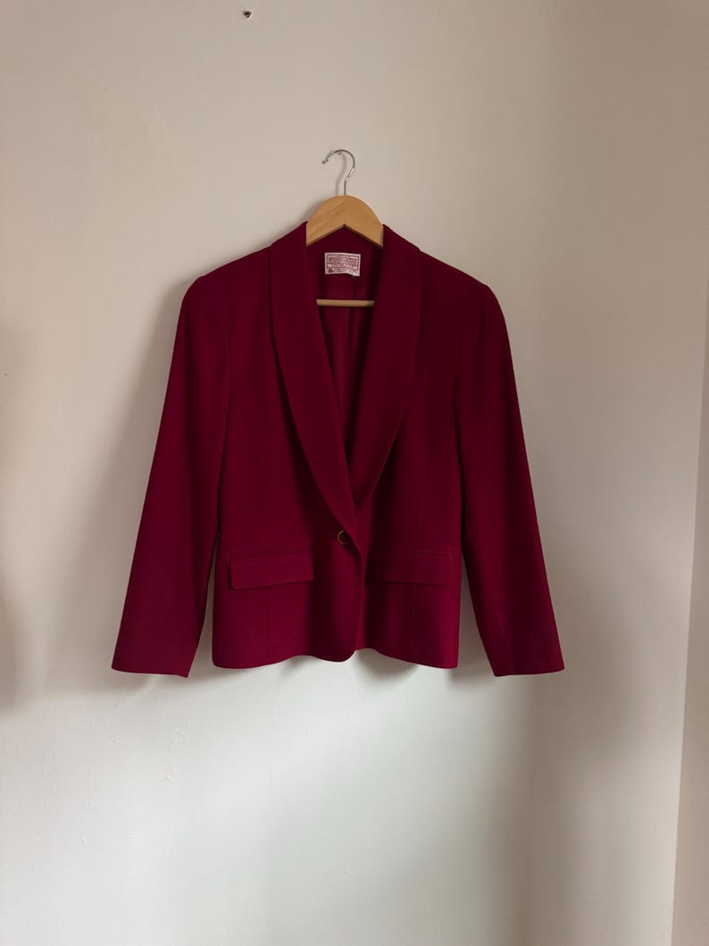 Pu&ograve; includere: Un blazer bordeaux con chiusura a un bottone e due tasche frontali. La giacca &egrave; appesa a una gruccia di legno contro un muro bianco. Il blazer ha un classico design a revers.