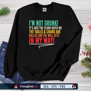 Puede incluir: Sudadera negra con el texto "I'M NOT DRUNK! IT'S JUST THE FLOOR HATES ME THE TABLES & CHAIRS ARE BULLIES AND THE WALL GETS IN MY WAY!". La sudadera se combina con pantalones cortos de mezclilla y adornos navideños.