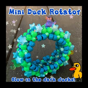 May include: A colorful beaded bracelet with the text "Mini Duck Rotator" and "Glow in the dark ducks!". The bracelet features blue, green, and clear beads, with star-shaped accents and a small duck figurine on top. A cartoon duck is in the lower right corner.