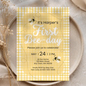 May include: A rectangular invitation card with a yellow and white gingham pattern. The text reads: It's Harper's First Bee-day, Please join us to celebrate, MAY 24 2 PM, AT OUR HOME 123 MAIN STREET ANY TOWN, WA 98208, KINDLY RSVP BY MAY 2ND.