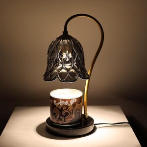 May include: A decorative candle warmer lamp with a dark grey, bell-shaped shade and a gold-coloured, curved arm. The base holds a candle with an ornate, brown and gold design. The lamp is on a white surface.