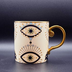 Op de afbeelding: Een witte keramische mok met een gouden handvat en een blauw en gouden oogontwerp. De mok heeft twee ogen aan de voorkant, elk met een blauwe iris en een gouden pupil.