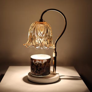 May include: A decorative table lamp with a gold-colored, curved metal frame. The lamp features a flower-shaped, amber-colored glass shade and a cylindrical base with a decorative pattern. The lamp is on a white surface.